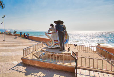 Men sitting on railing by sea against sky