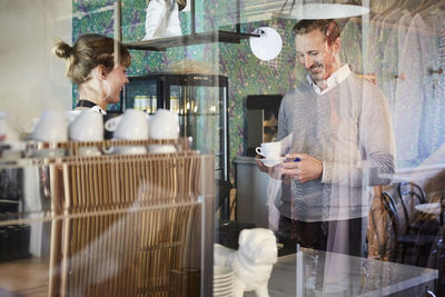 Owner looking at customer having coffee at checkout counter seen through glass in cafe