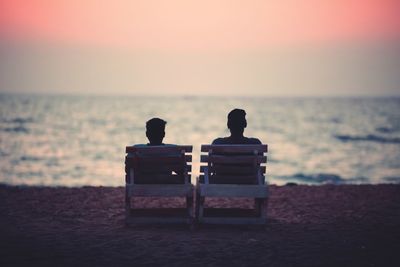 Rear view of couple sitting on chairs at beach against sky during sunset