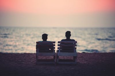 Rear view of man sitting on shore against sea during sunset