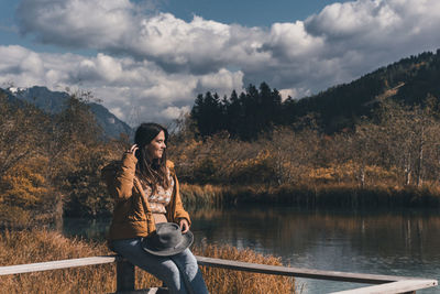 Young woman sitting on fence on shore of lake in autumn
