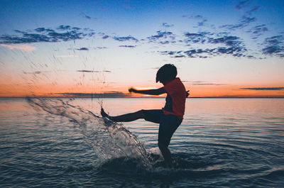Boy splashing water in sea against sky during sunset