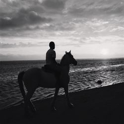 Man riding horse at beach against cloudy sky during sunset