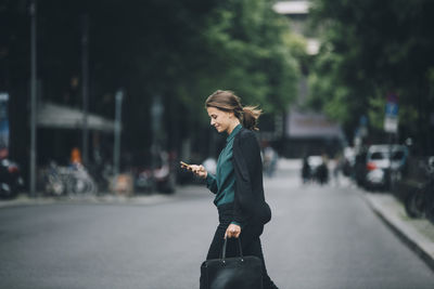 Confident businesswoman using smart phone while crossing street in city