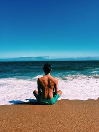 Rear view of shirtless man sitting on shore at beach against blue sky