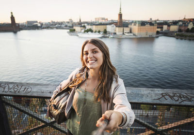 Portrait of happy female tourist holding monopod on bridge by river