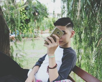 Portrait of young woman holding camera against trees