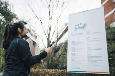 Female worker carrying box and document with smart phone looking at billboard