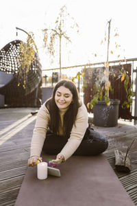 Smiling woman burning candle while sitting on exercise mat at terrace