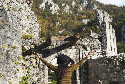 Rear view of woman in autumn clothes standing in old castle ruin, arms outstretched.