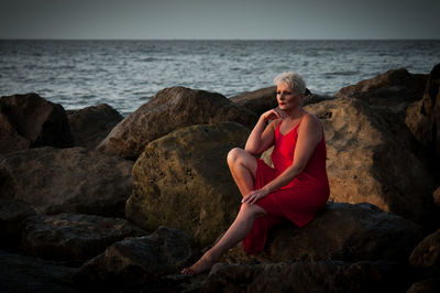 Young woman sitting on rock at sea shore against sky