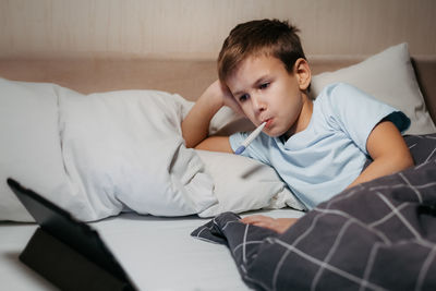 Sad child laying in bed with thermometer in his mouth and look at tablet. sick boy watching cartoons