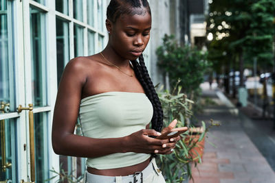 Young african american woman with long braids wearing stylish tube top and browsing smartphone on city street