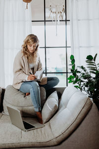 Woman writing in book while sitting by laptop on sofa at home