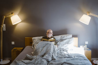 Senior man spending leisure time while reading book on bed at home