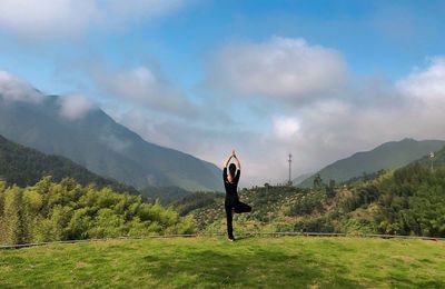 Full length rear view of woman doing yoga while standing on grass against mountains and sky