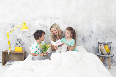 Smiling woman holding greeting card while sitting with children on bed at home