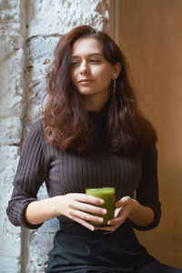 Smiling woman having drink while sitting by wall