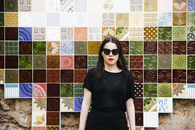 Portrait of young woman wearing sunglasses while standing against tiled wall