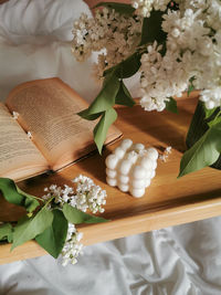 Open book, bubble cube soy candle and bunch of white lilac flowers on folding wooden table in bed.
