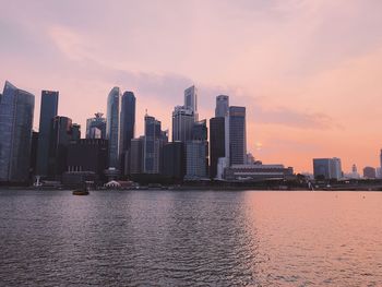 Modern buildings by river against sky during sunset