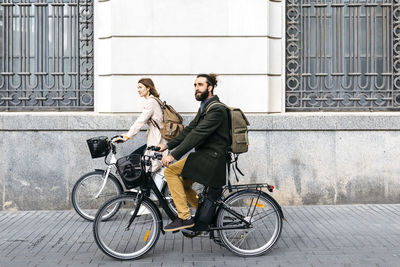 Couple riding e-bikes in the city passing a building