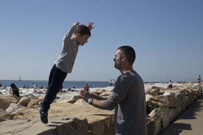 Side view of man catching son jumping at beach against clear blue sky