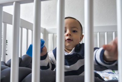 Curious cute little baby sitting in crib and looking at camera in light room in daytime