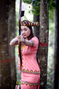 Portrait of young woman holding bow and arrow standing by tree outdoors