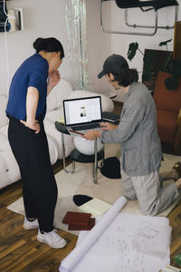 Multiracial male and female architects discussing laptop by blueprint at home office