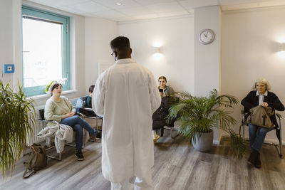 Rear view of male doctor talking to patients sitting in waiting room at hospital