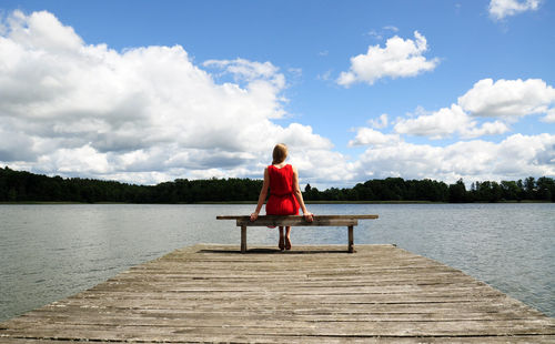 Rear view of woman sitting on pier over lake against cloudy sky