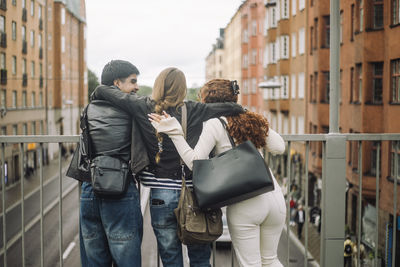 Rear view of teenage girl standing with arms around male and female friends near railing in city