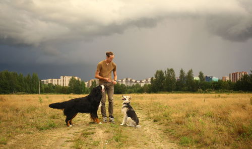 Man playing with dogs while standing on grassy land against cloudy sky