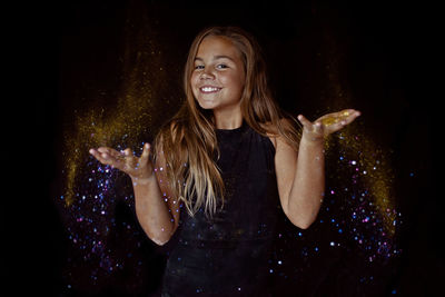 Portrait of smiling girl throwing glitter against black background