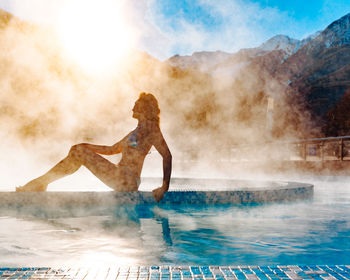 Slender model girl sits in a swimsuit in a winter outdoor pool in a steam against a sunny blue sky