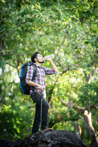 Young man drinking water while standing on rock by trees in forest