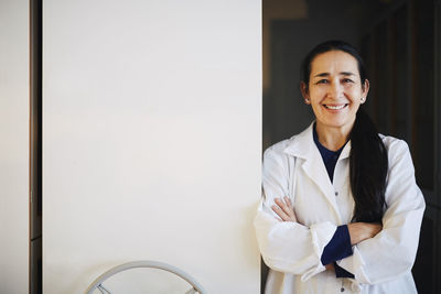 Portrait of smiling mature female teacher standing with arms crossed against storage cabinets in university