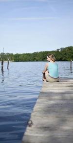 Rear view of woman sitting on jetty over lake against sky