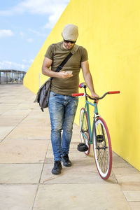 Man with bicycle using mobile phone while standing on footpath by yellow wall