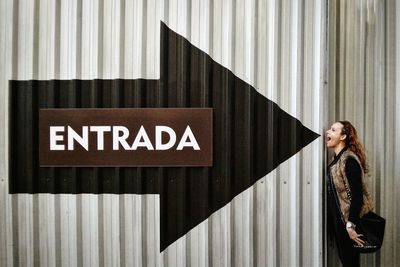 Side view of woman with mouth open standing by arrow sign on corrugated wall