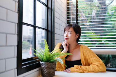 Young woman looking away while sitting on window at home