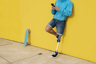 Young disabled athlete using mobile phone while standing against yellow wall