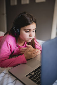 Focused little girl in headphones lying on bed and watching online video on laptop while spending free time at home