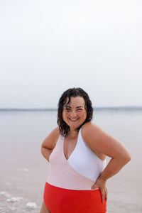 Positive curvy female in swimsuit standing with hands on waist on beach near pink pond and looking at camera while enjoying summer holiday