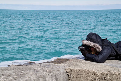 Woman photographing while lying on retaining wall by sea