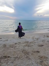 Rear view of woman standing on sand at beach against cloudy sky during sunset