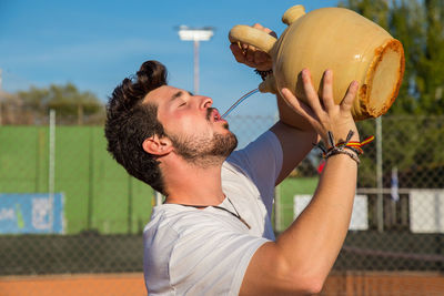 Young man drinking water from pot at court during sunny day