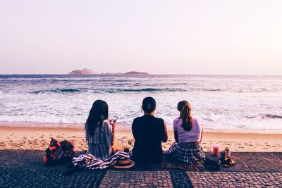 Rear view of female friends relaxing at beach against sky