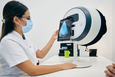 Side view of female cosmetologist using modern facial diagnostics machine for analyzing skin of client in beauty clinic